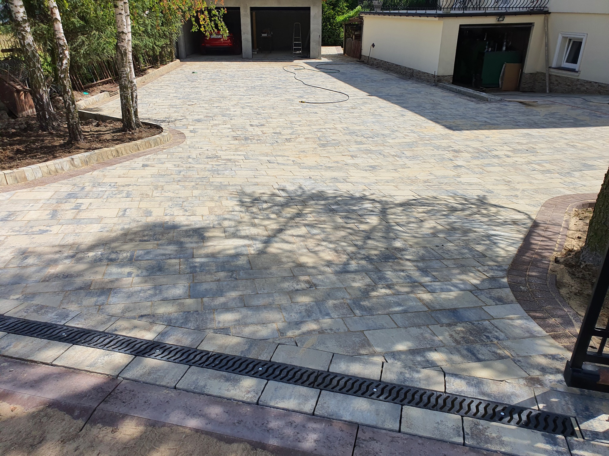 Szeroki kadr na podjazd z kostki brukowej w odcieniach szarości i beżu, z widocznym drenażem liniowym i obrzeżami. W tle budynek z garażami i drzewa.