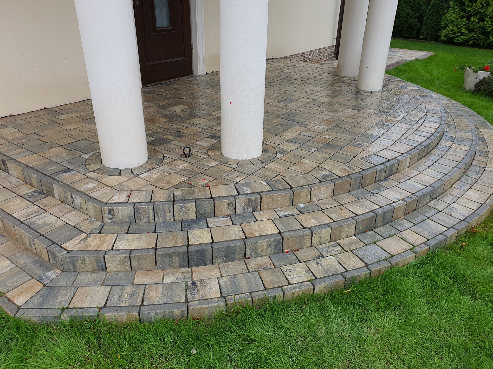 Schody wejściowe z kostki brukowej w odcieniach szarości i beżu, otoczone zielonym trawnikiem, z białymi kolumnami podtrzymującymi zadaszenie.