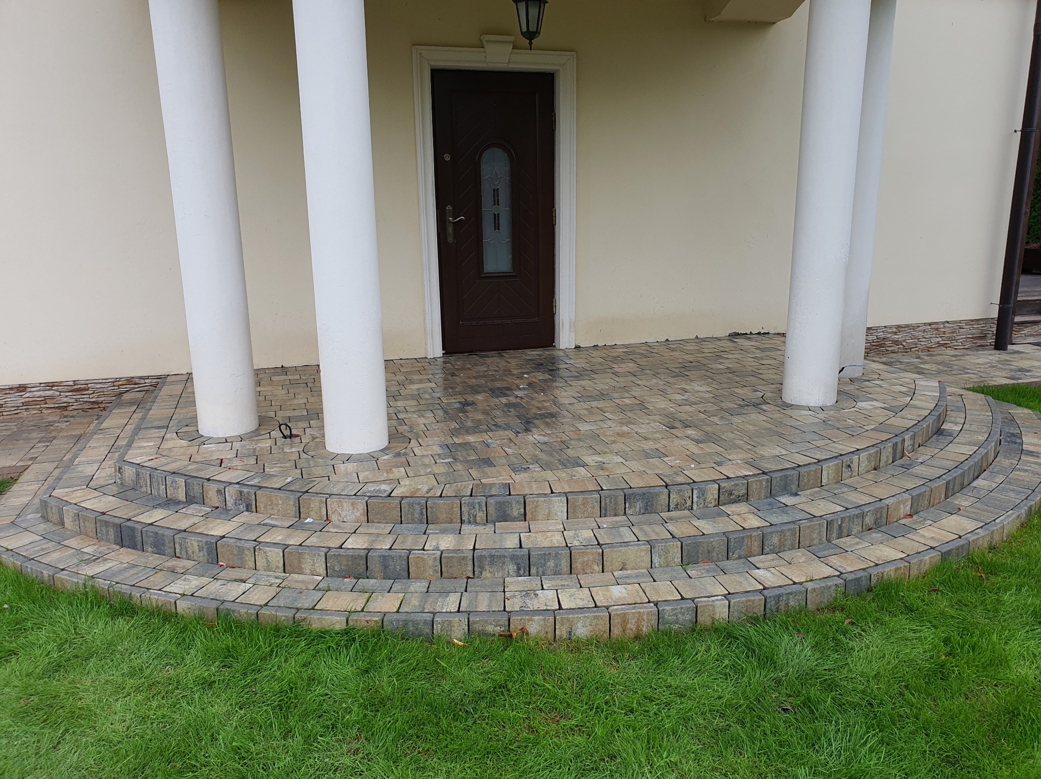 Półokrągłe schody z kostki brukowej prowadzące do wejścia z kolumnami. Kostka w odcieniach szarości i beżu, otoczenie zielony trawnik. Widok na elewację budynku.