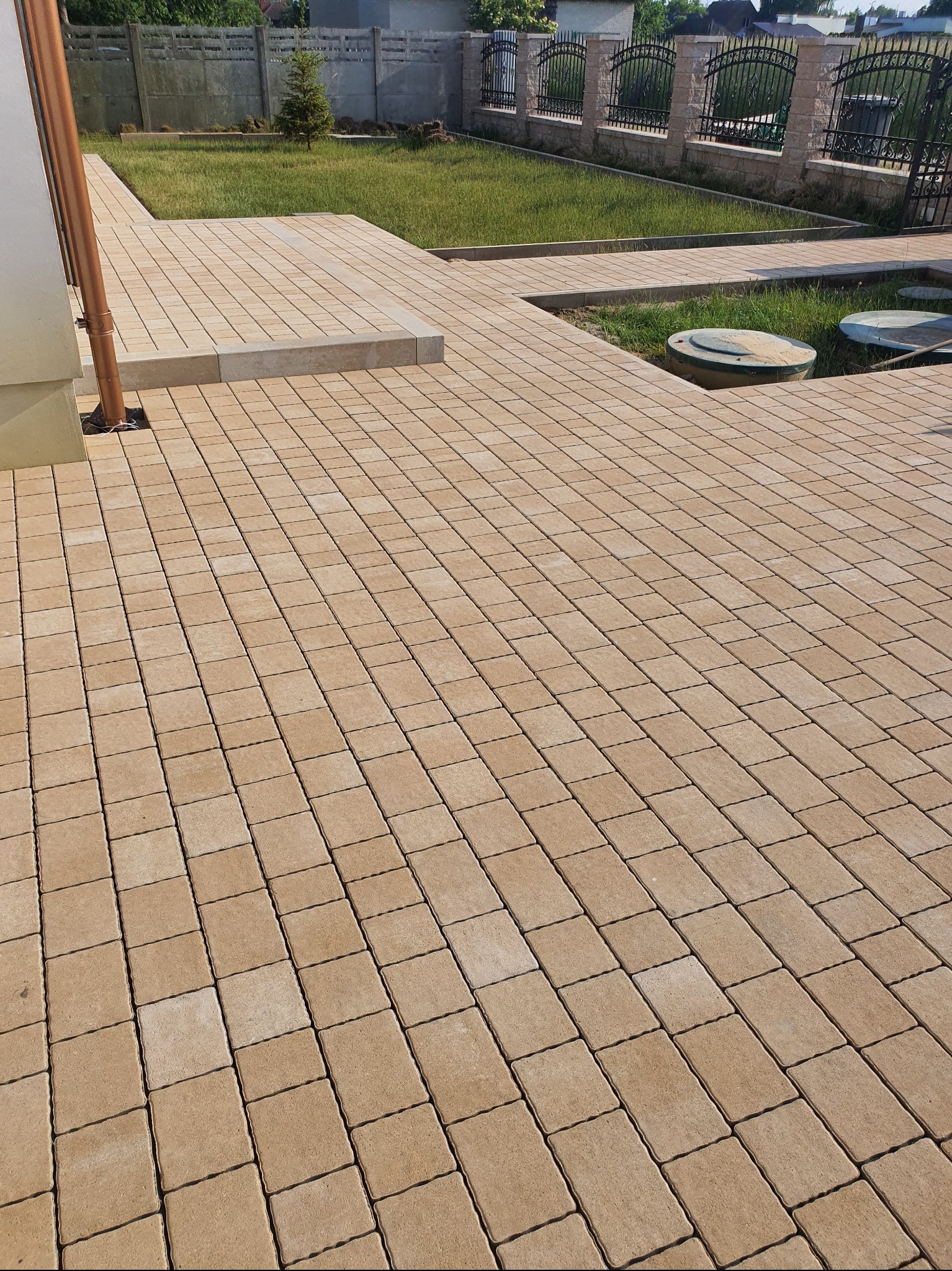 Beżowa kostka brukowa ułożona na posesji, widoczne studzienki kanalizacyjne, trawnik i ogrodzenie z metalowymi przęsłami. Ujęcie z niskiej perspektywy.