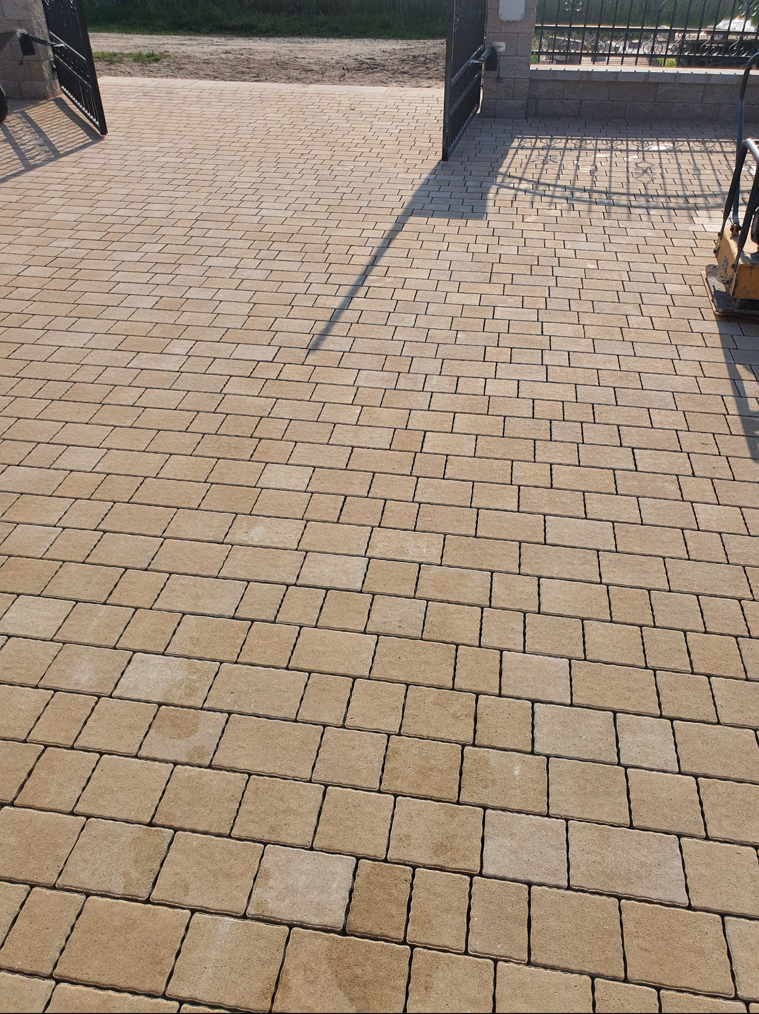 Beżowa kostka brukowa ułożona na podjeździe, widoczna brama wjazdowa i ubijarka wibracyjna. Regularny wzór kostki, ciepłe oświetlenie słoneczne.