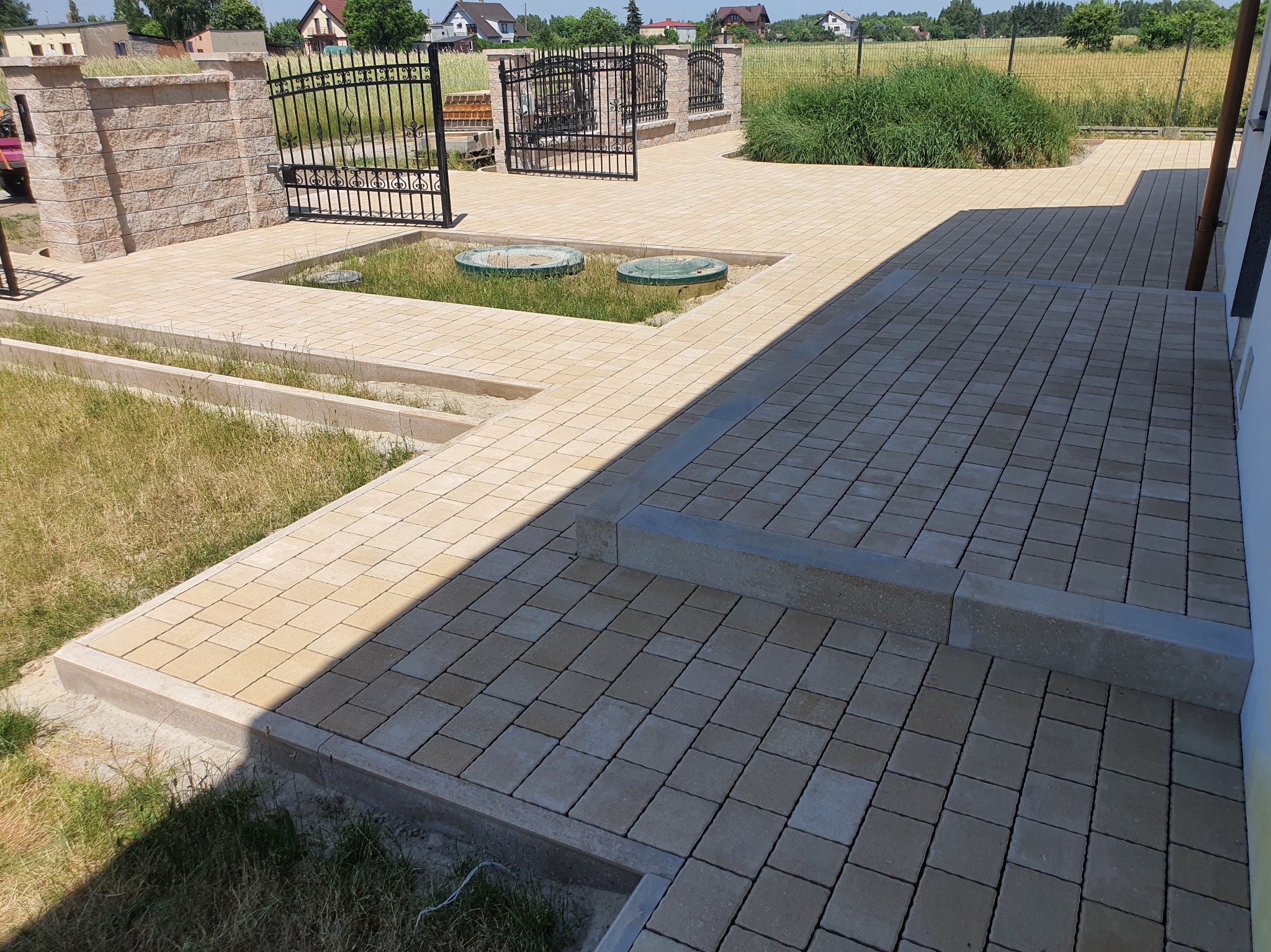 Jasna kostka brukowa ułożona na posesji w Zduńskiej Woli. Widoczne studzienki kanalizacyjne i brama wjazdowa. Estetyczne wykończenie terenu.