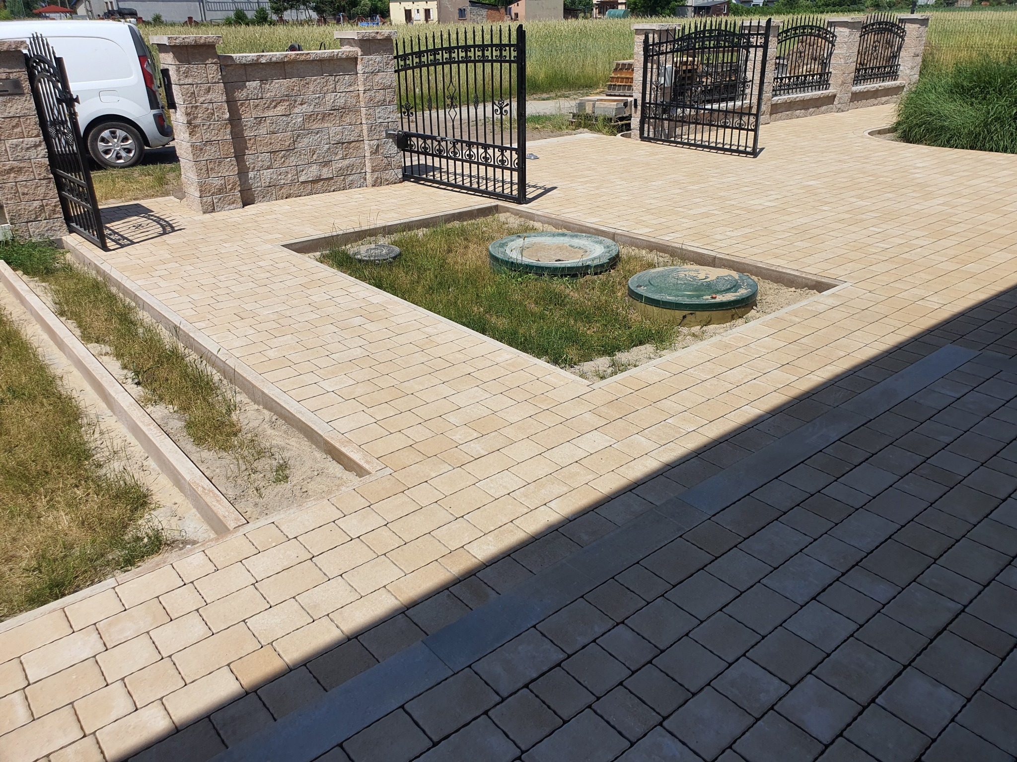 Jasna kostka brukowa tworzy ścieżkę i podjazd, z czarnymi, kutymi bramami i ogrodzeniem w tle. Widoczne studzienki kanalizacyjne w trawniku i fragment białego samochodu.