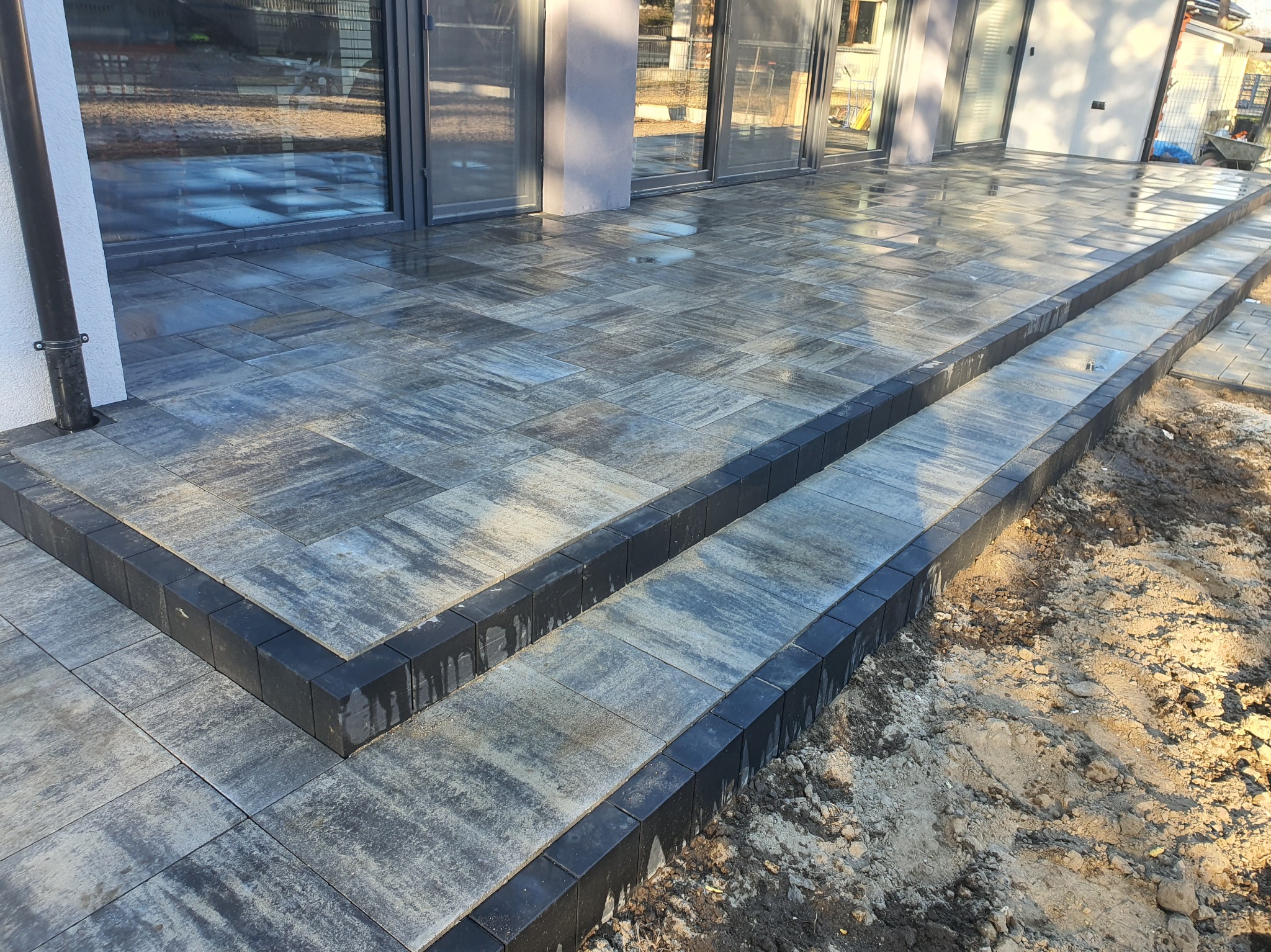 Ułożona, szara kostka brukowa tworząca schody i taras przy nowoczesnym domu z dużymi oknami. Ciemne obrzeża kostki podkreślają krawędzie. Widoczny fragment gruntu.