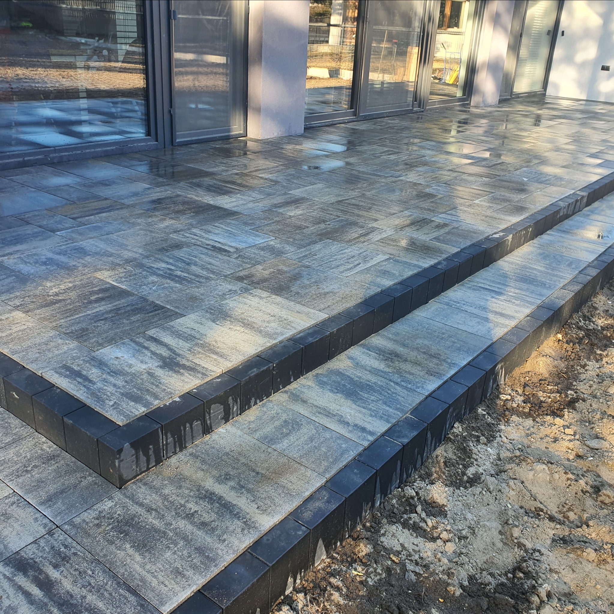 Ułożona kostka brukowa w odcieniach szarości tworząca schody i taras przy budynku z dużymi oknami. Krawężniki z ciemnej kostki. Widoczne ślady po deszczu.
