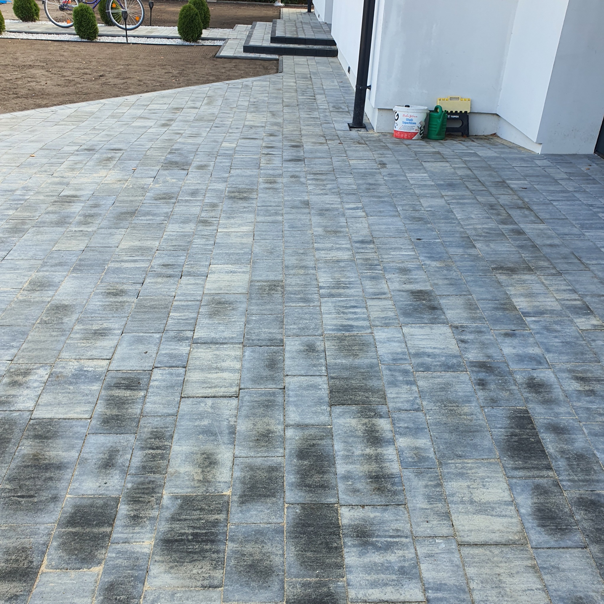 Szara kostka brukowa ułożona na podjeździe, widok z perspektywy, z elementami zieleni i fragmentem budynku w tle. Widoczne ślady użytkowania na kostce.