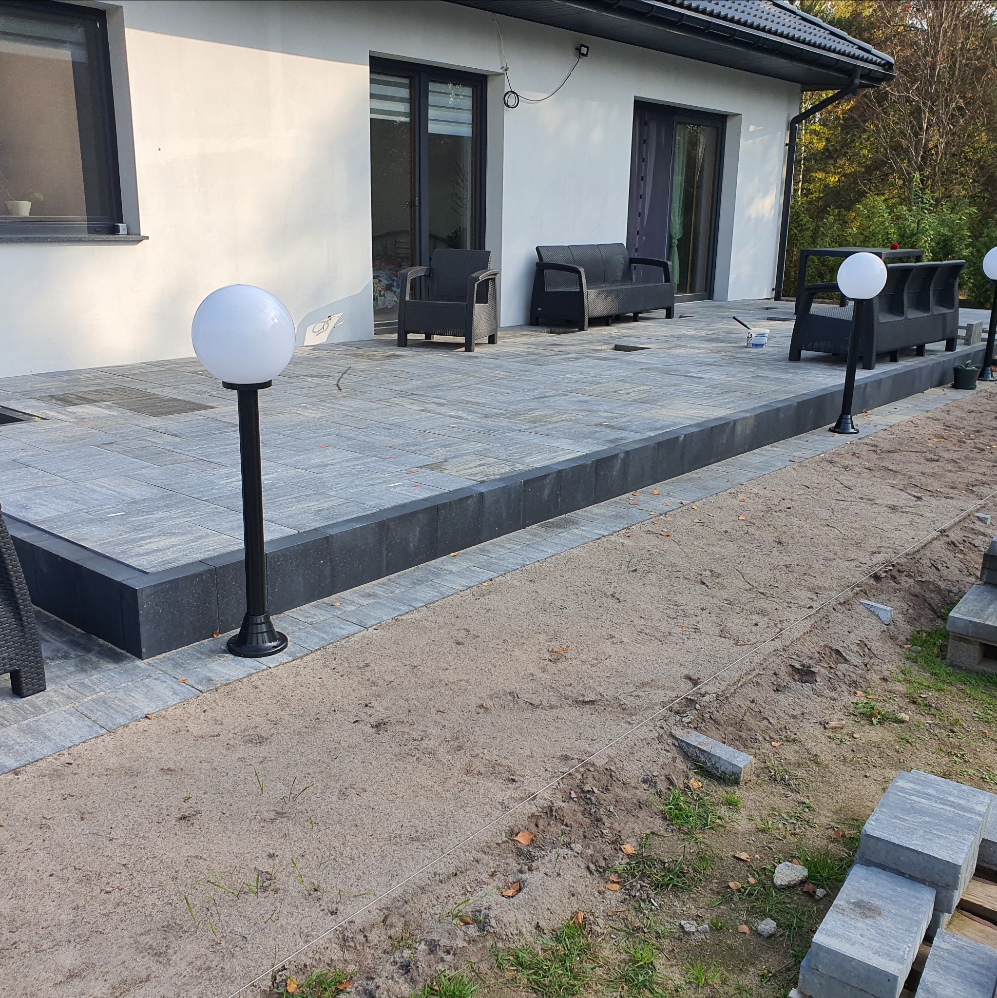 Nowoczesny taras z szarej kostki brukowej, oświetlony kulistymi lampami. Widoczne meble ogrodowe i przygotowany teren pod dalsze prace.