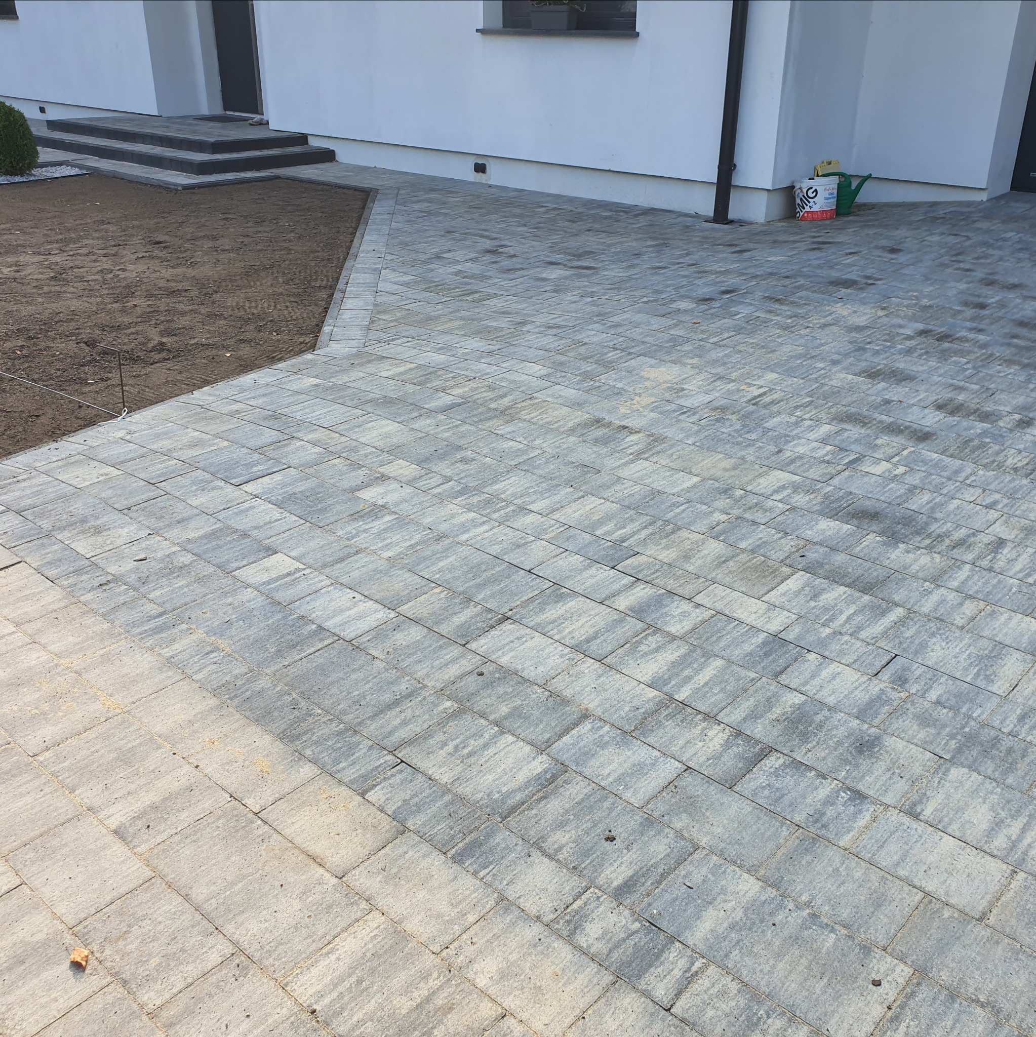 Ułożona szara kostka brukowa na podjeździe domu jednorodzinnego. Widoczny fragment rabaty z ziemią i schody wejściowe. W tle biała ściana budynku.