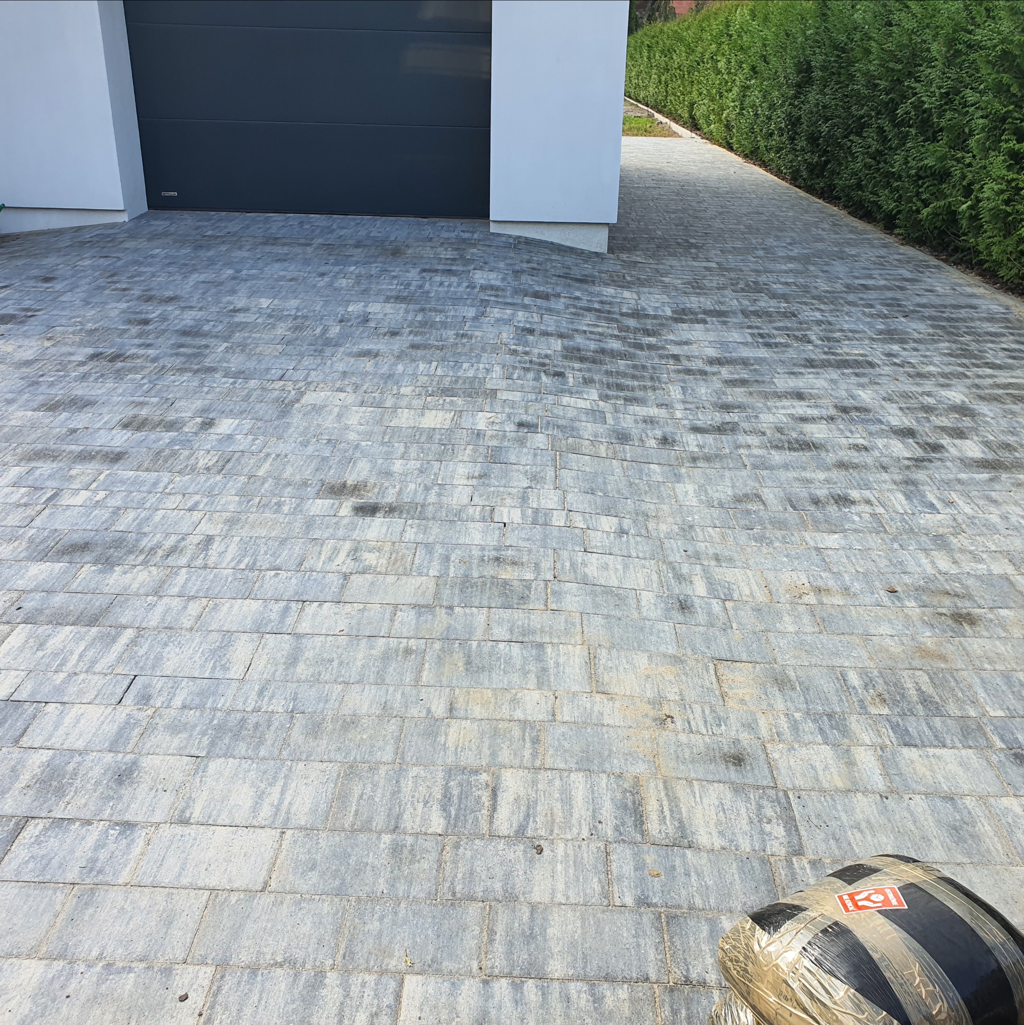 Ułożona kostka brukowa w odcieniach szarości prowadząca do garażu i wzdłuż żywopłotu. W rogu widoczny pakiet kostki brukowej.