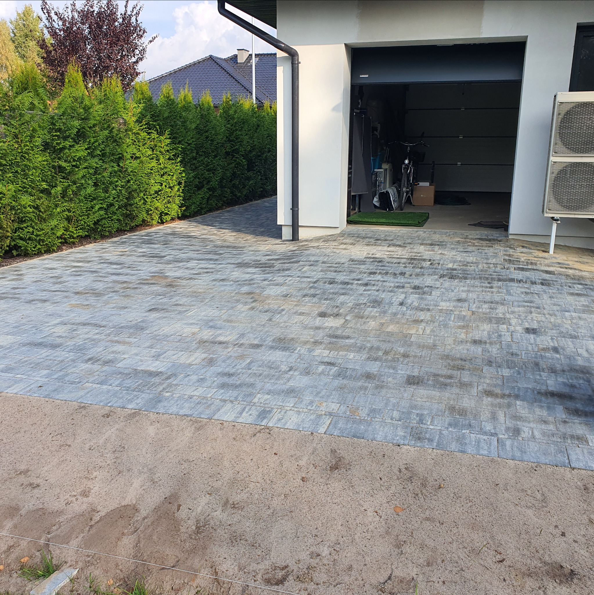 Ułożona, szara kostka brukowa prowadząca do garażu, obok zadbane tuje. Część terenu jeszcze nie wykończona. Widok na dom z ciemnym dachem.