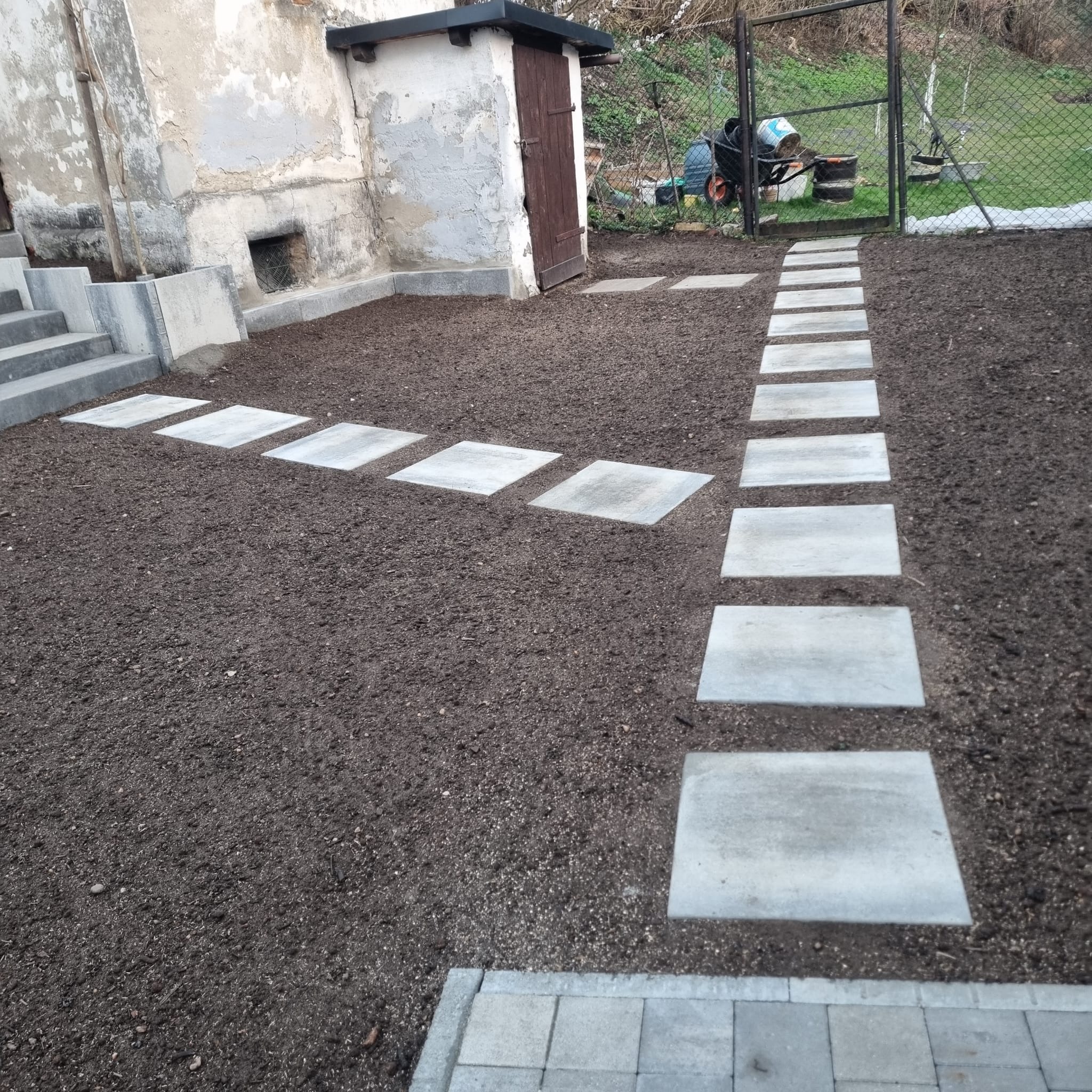 Ścieżka z kwadratowych płyt betonowych w świeżo przygotowanym gruncie, prowadząca od schodów wzdłuż budynku gospodarczego, z widocznym ogrodem w tle.