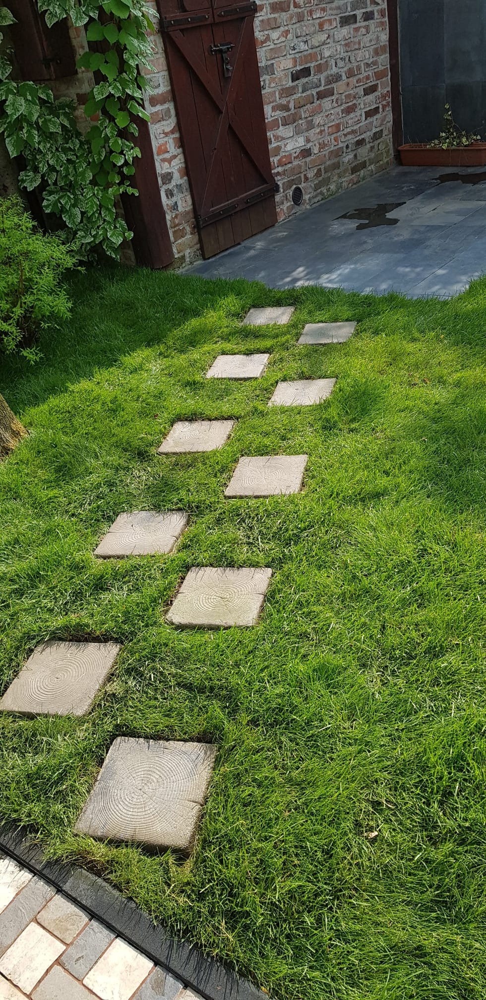 Ścieżka z kwadratowych płyt drewnianych w trawie, prowadząca do budynku z cegły i drewnianymi drzwiami. Układ nieregularny, trawa świeżo skoszona.