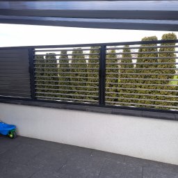 Usługi ślusarskie Paweł Drzazga - Nowoczesna zabudowa balkonu z szarymi, poziomymi lamelami i metalową ramą, zapewniająca prywatność i estetyczny wygląd. W tle widoczne drzewa.