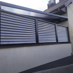 Usługi ślusarskie Paweł Drzazga - Nowoczesna, szara pergola z lamelami na balkonie, z widocznym klimatyzatorem na dachu budynku w tle. Minimalistyczny design, poziome linie, stonowana kolorystyka.