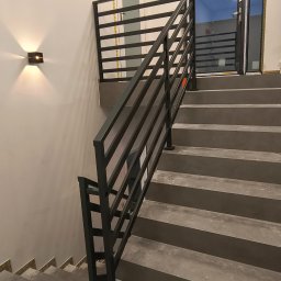 Usługi ślusarskie Paweł Drzazga - Nowoczesne schody z szarą betonową konstrukcją i czarną, poziomą balustradą metalową. Minimalistyczny design, oświetlenie ścienne, widok z dołu na górę.