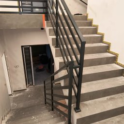Usługi ślusarskie Paweł Drzazga - Nowoczesne schody betonowe z czarną, metalową balustradą o minimalistycznym designie. Widoczne zabezpieczenia na krawędziach stopni. W tle drzwi i fragment pomieszczenia.