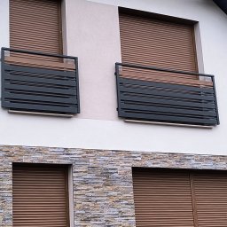 Usługi ślusarskie Paweł Drzazga - Nowoczesna, grafitowa balustrada balkonowa z poziomymi panelami, zamontowana na balkonie z brązowymi roletami zewnętrznymi. Elewacja budynku z kamienną okładziną.