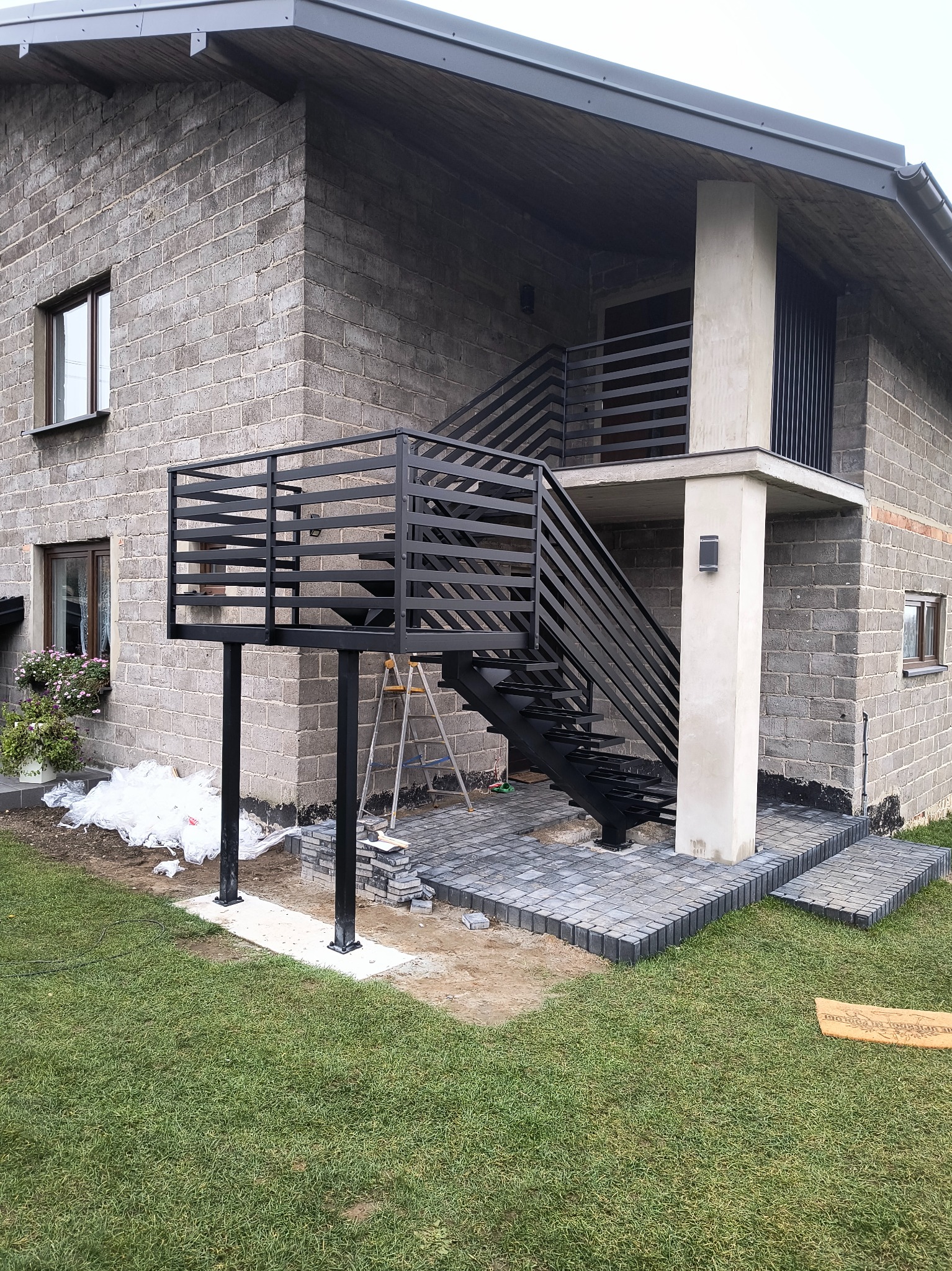 Nowoczesne, czarne schody metalowe z balustradą prowadzące na balkon w domu z szarej cegły. Widoczne kostki brukowe i trawnik.