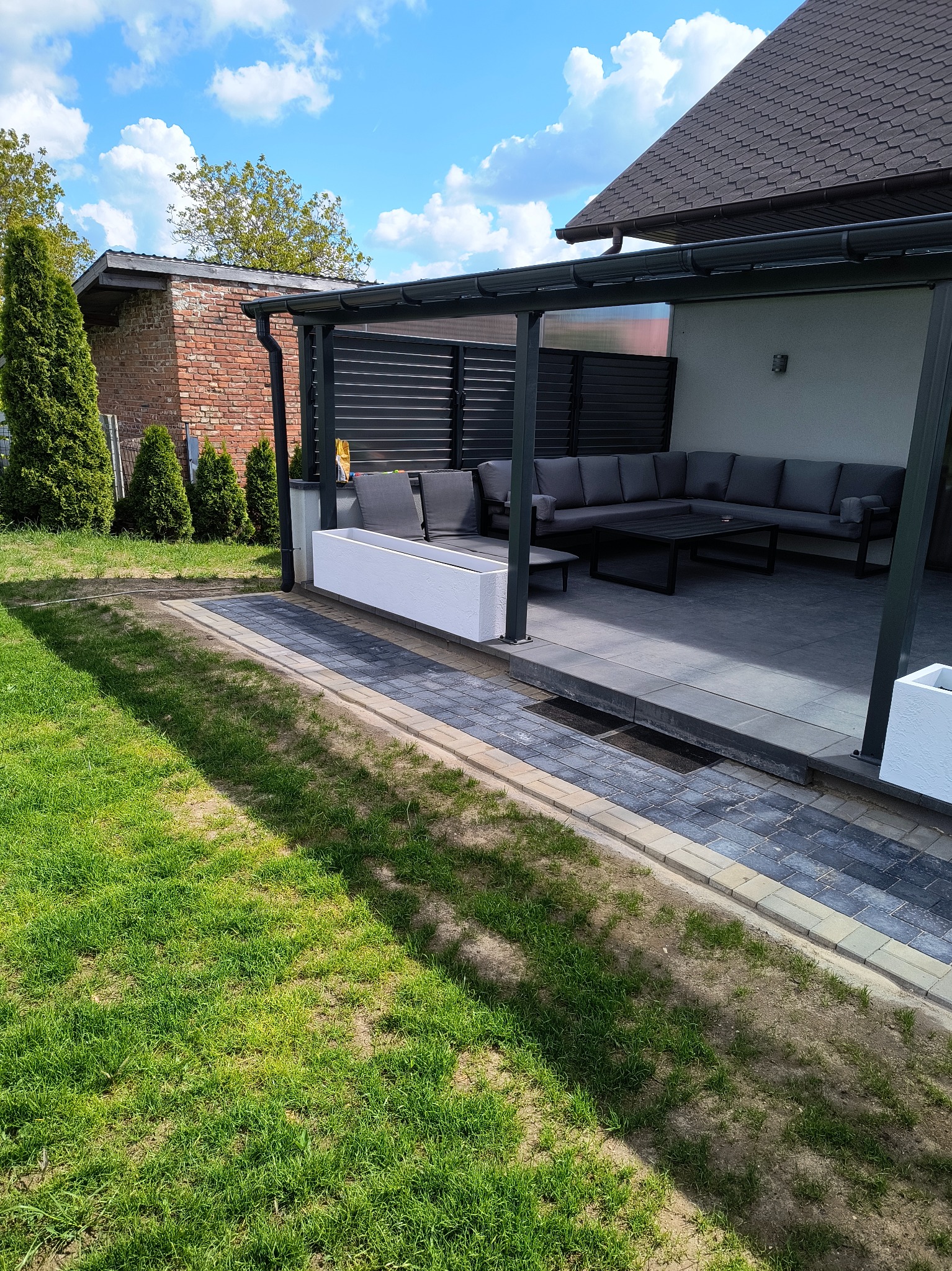 Nowoczesna pergola z szarym metalowym stelażem i poziomymi panelami, z wygodnym narożnikiem i stolikiem kawowym na wyłożonym kostką tarasie, otoczona zielenią.