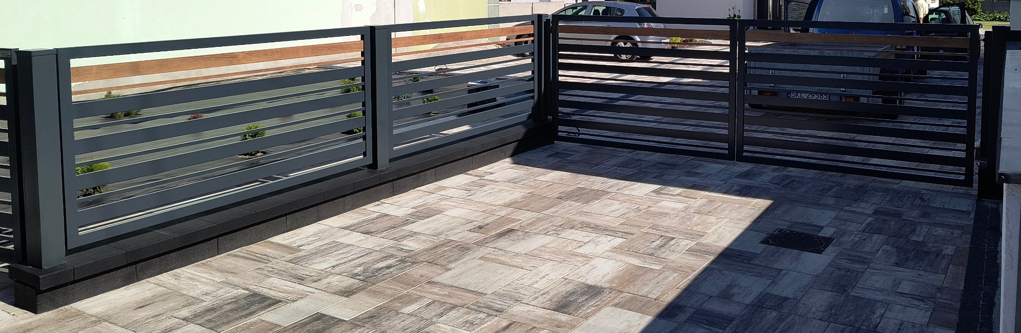Nowoczesne ogrodzenie posesji z poziomych, szarych paneli metalowych z drewnianymi akcentami, osadzone na niskim, ciemnym murku. Widok na kostkę brukową i zaparkowane auta.