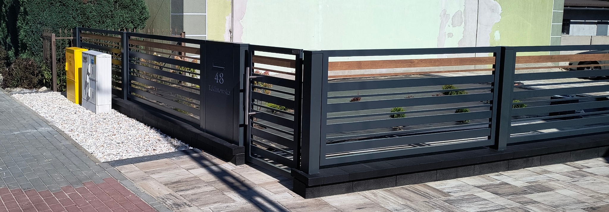 Nowoczesna brama wjazdowa z poziomymi, grafitowymi przęsłami i drewnianymi akcentami. Skrzynka na listy z numerem domu '48 Teklinowska'.