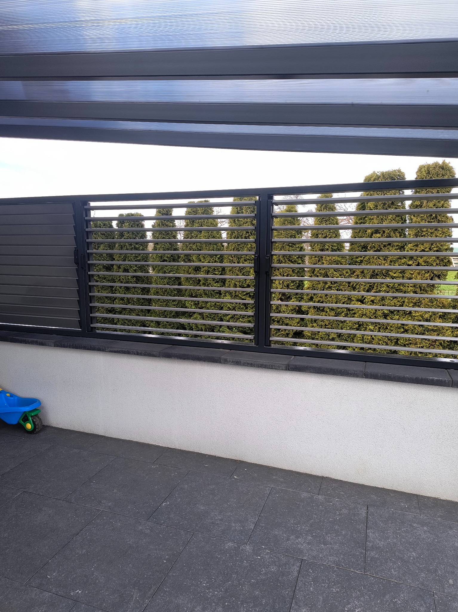 Nowoczesna zabudowa balkonu z szarymi, poziomymi lamelami i metalową ramą, zapewniająca prywatność i estetyczny wygląd. W tle widoczne drzewa.