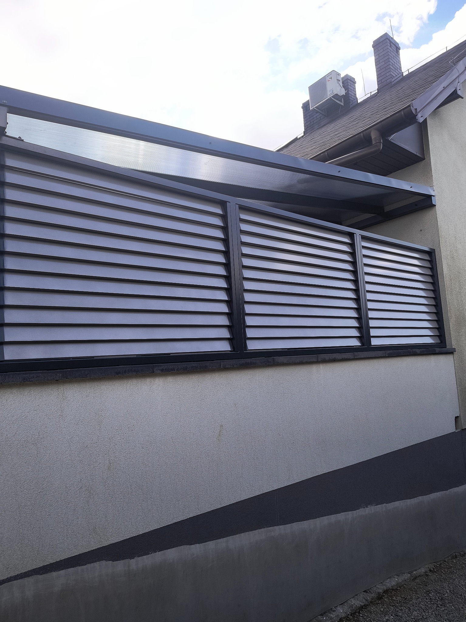 Nowoczesna, szara pergola z lamelami na balkonie, z widocznym klimatyzatorem na dachu budynku w tle. Minimalistyczny design, poziome linie, stonowana kolorystyka.