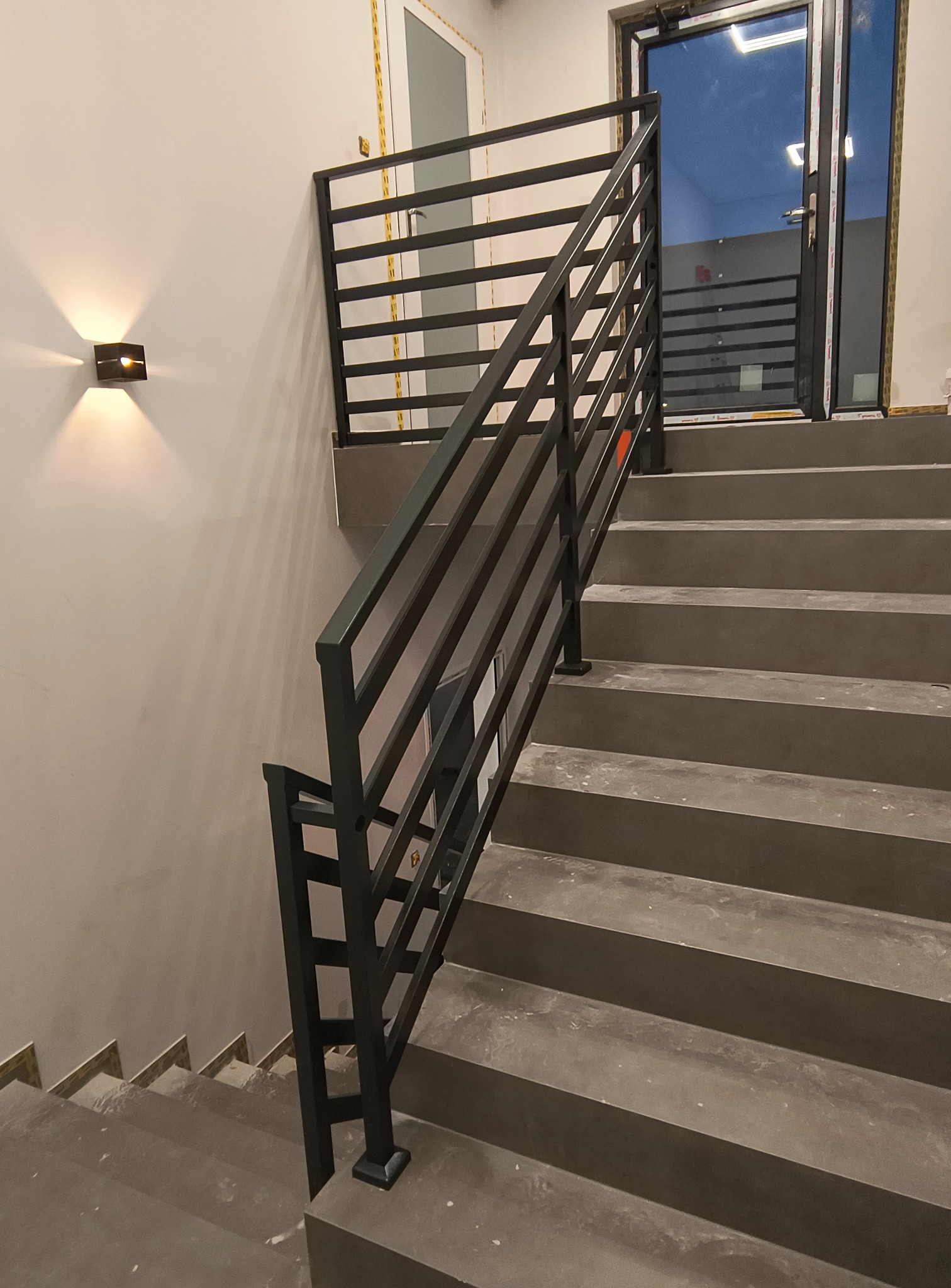 Nowoczesne schody z szarą betonową konstrukcją i czarną, poziomą balustradą metalową. Minimalistyczny design, oświetlenie ścienne, widok z dołu na górę.