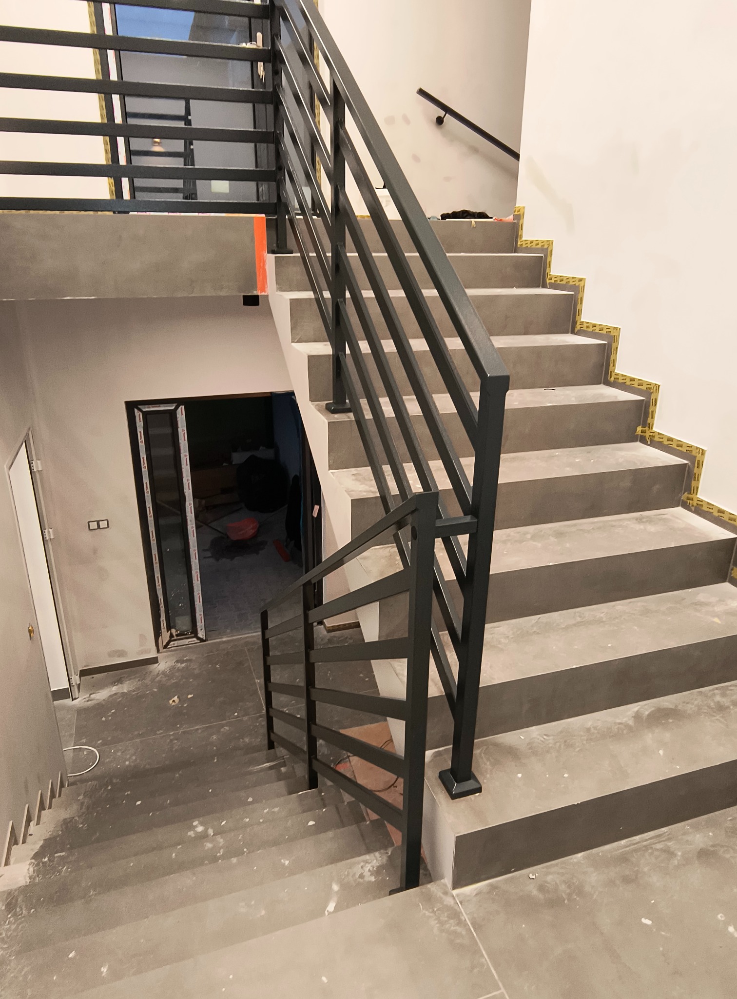 Nowoczesne schody betonowe z czarną, metalową balustradą o minimalistycznym designie. Widoczne zabezpieczenia na krawędziach stopni. W tle drzwi i fragment pomieszczenia.
