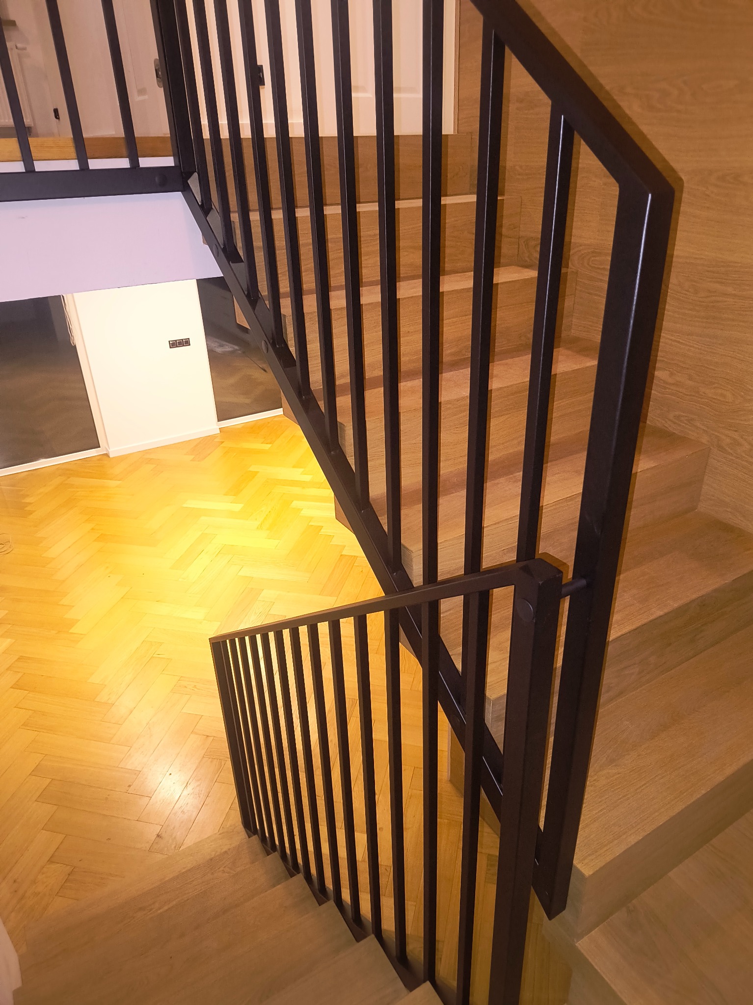 Nowoczesne schody drewniane z czarną, metalową balustradą. Widok z góry na parkiet w jodełkę i minimalistyczne wykończenie wnętrza.