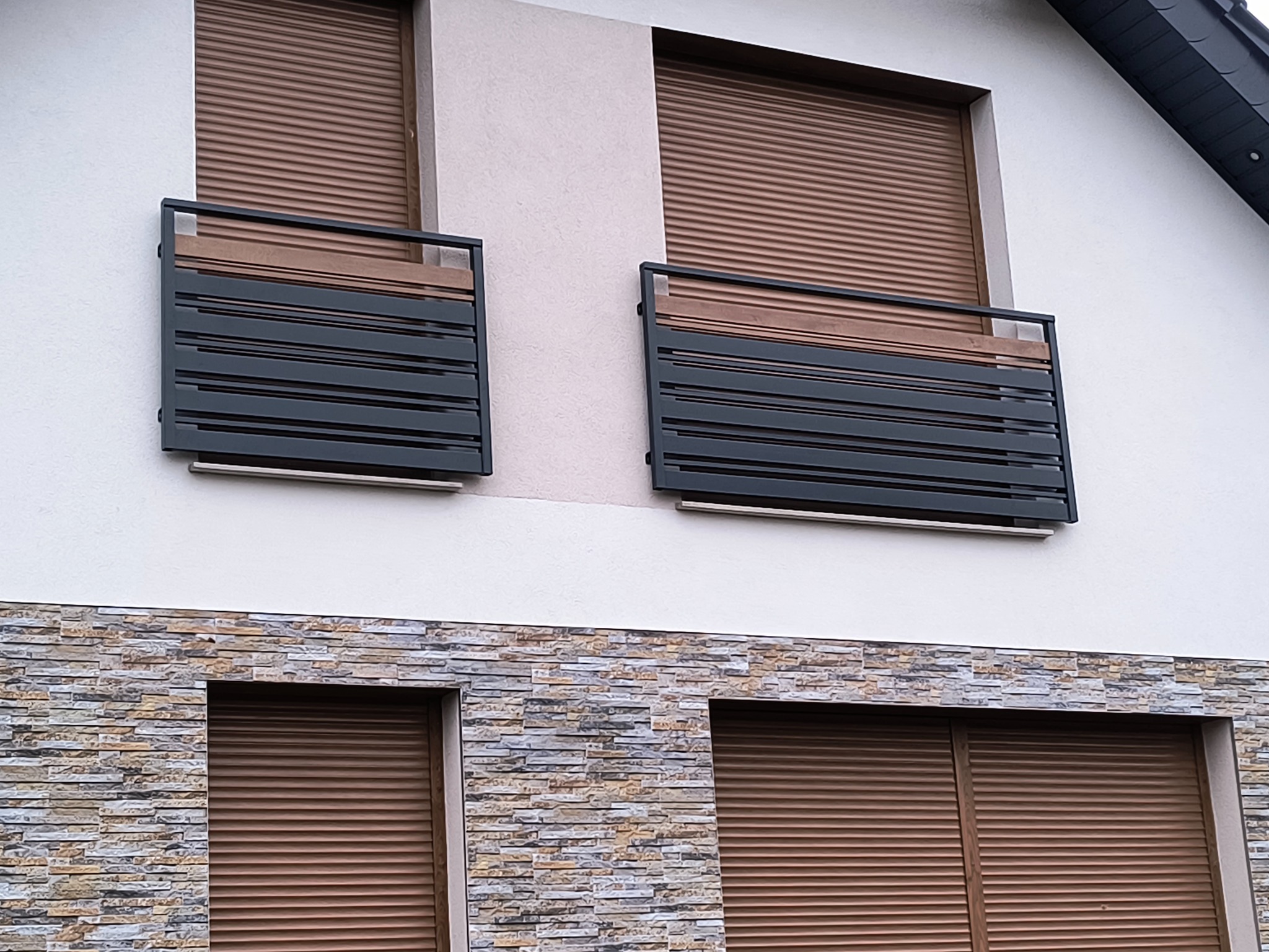 Nowoczesna, grafitowa balustrada balkonowa z poziomymi panelami, zamontowana na balkonie z brązowymi roletami zewnętrznymi. Elewacja budynku z kamienną okładziną.