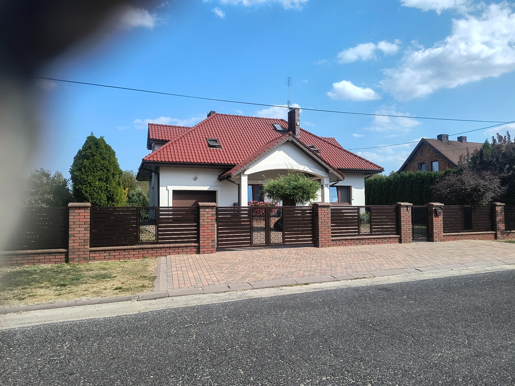 Nowoczesne ogrodzenie z brązowych, poziomych paneli i ceglanych słupków, z dekoracyjnymi elementami ażurowymi przy bramie wejściowej do domu.