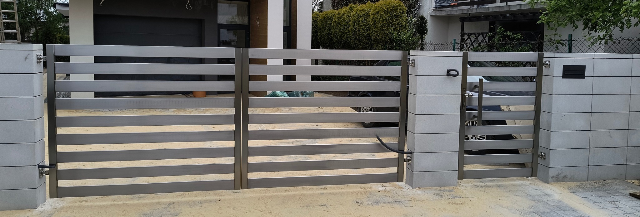 Nowoczesna brama wjazdowa z poziomych, metalowych profili, osadzona na betonowych słupkach. Widoczna skrzynka na listy i dom w tle.