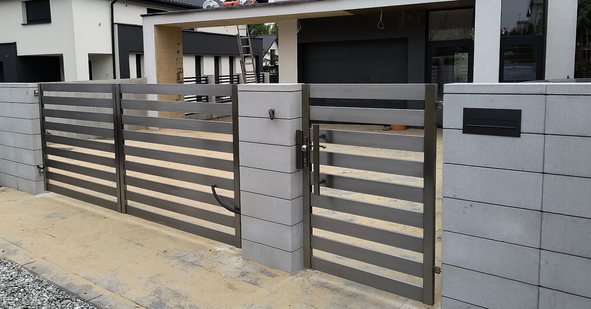 Nowoczesna brama wjazdowa z poziomych, metalowych profili, osadzona na betonowych słupkach z czarną skrzynką na listy. Widok na dom z garażem.
