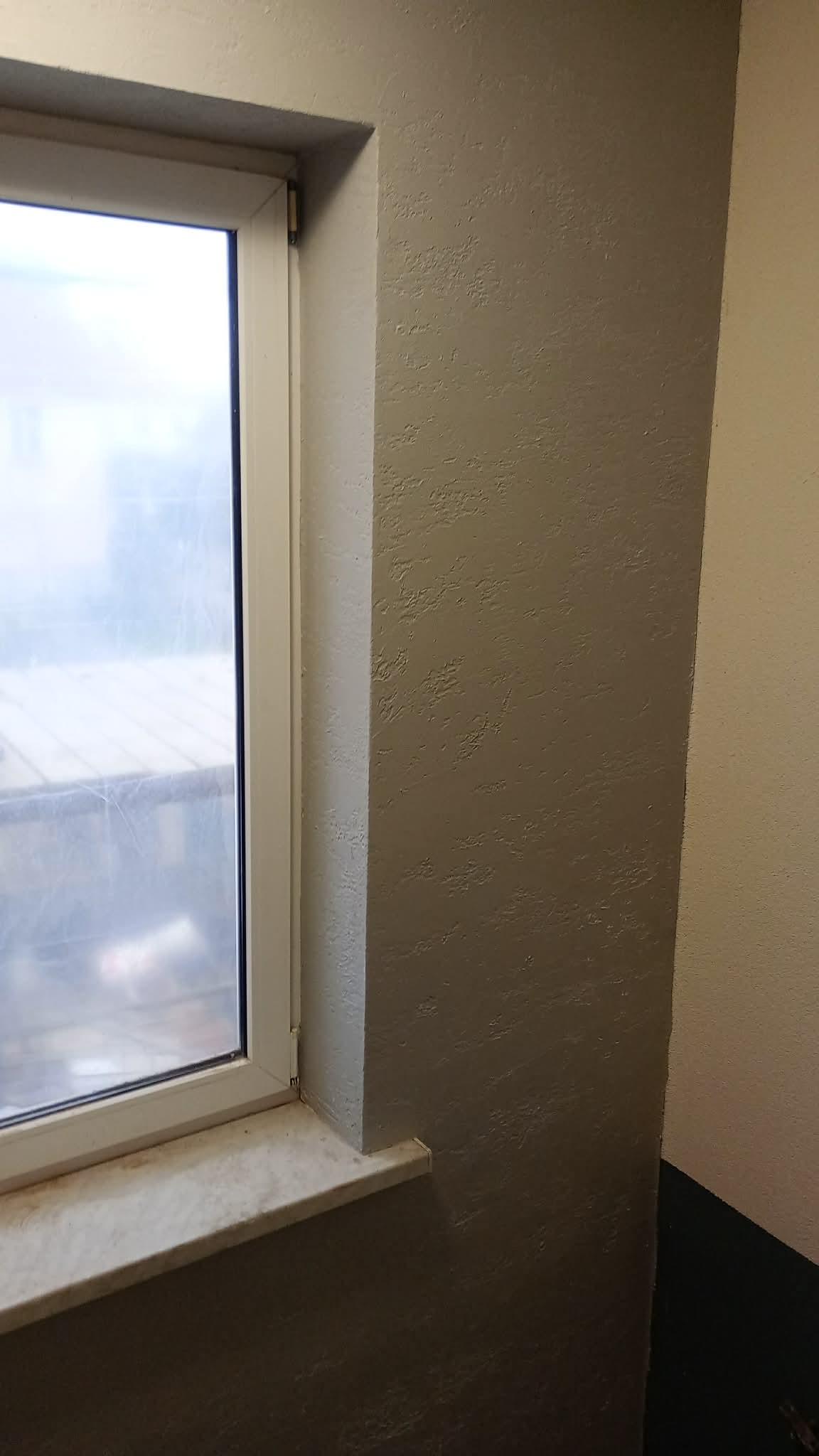 Szara ściana z fakturą przy oknie z białymi ramami i marmurowym parapetem. Widoczne wykończenie narożnika ściany. Teksturowana powierzchnia.