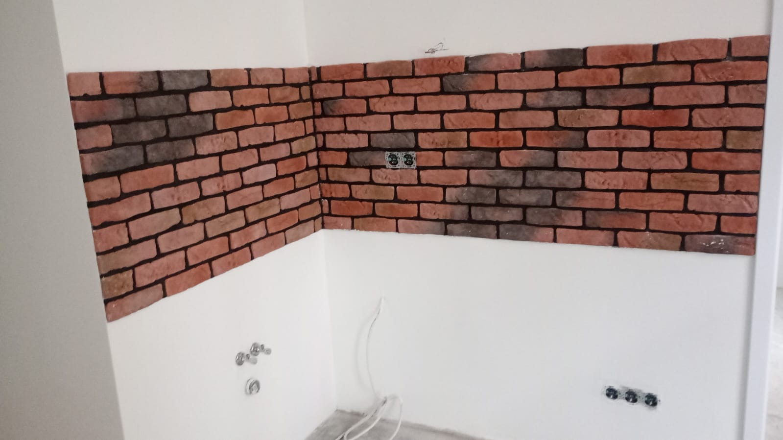 Ściana wykończona czerwoną cegłą dekoracyjną z widocznymi gniazdkami elektrycznymi i instalacją wodną. Część ściany pomalowana na biało.