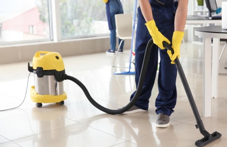 Pracownik w niebieskim kombinezonie i żółtych rękawicach odkurza jasną, błyszczącą podłogę w biurze żółtym odkurzaczem przemysłowym. W tle okno i inna osoba.