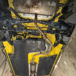 Patryk Jawor - Żółte podwozie auta po konserwacji antykorozyjnej, widok od spodu. Elementy zawieszenia i układu wydechowego w kolorze czarnym. Widoczne ślady po konserwacji.