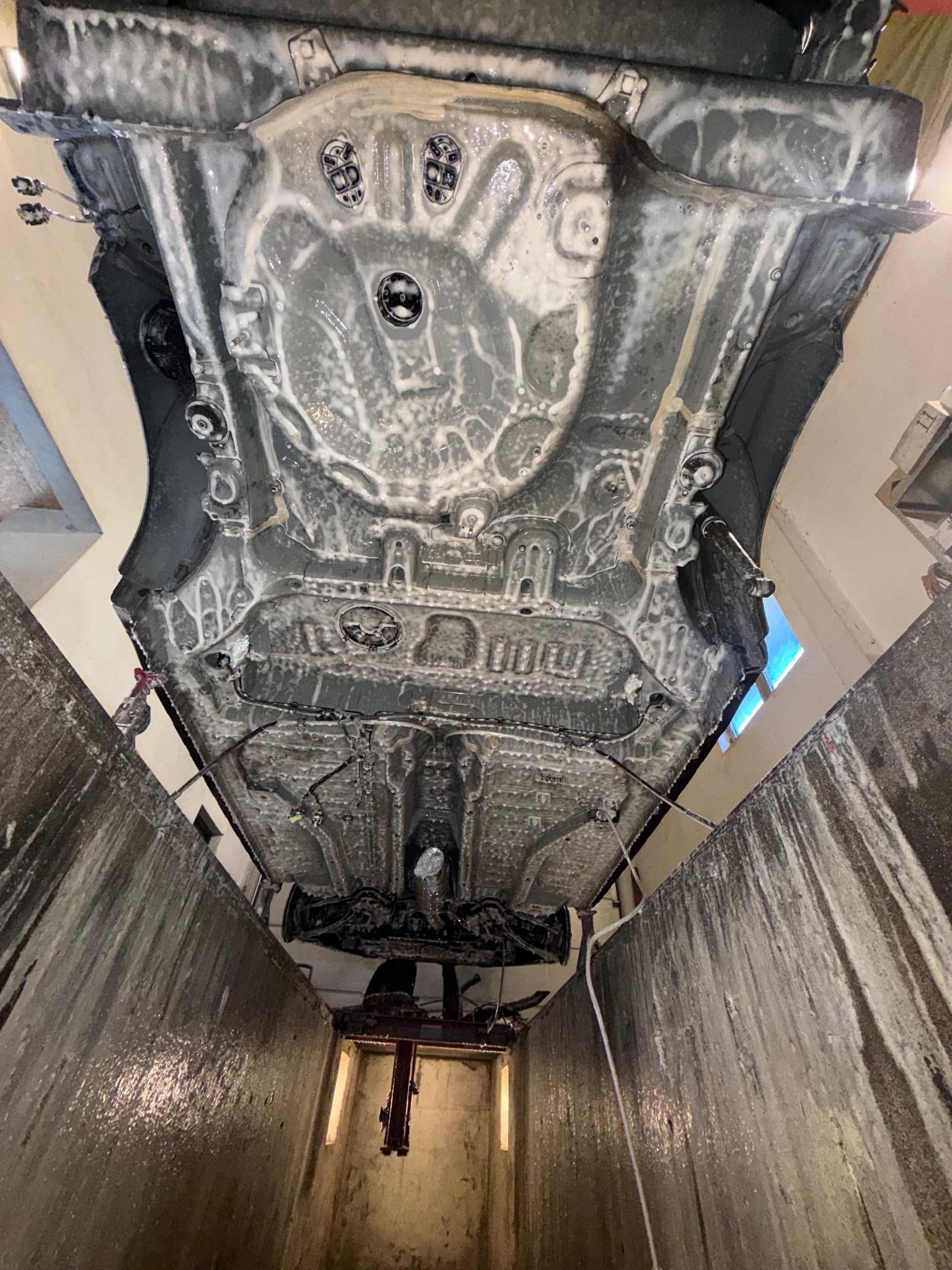 Podwozie samochodu pokryte pianą czyszczącą, widoczne z kanału warsztatowego. Specyficzny kąt ujęcia podkreśla detale konstrukcji i proces mycia.