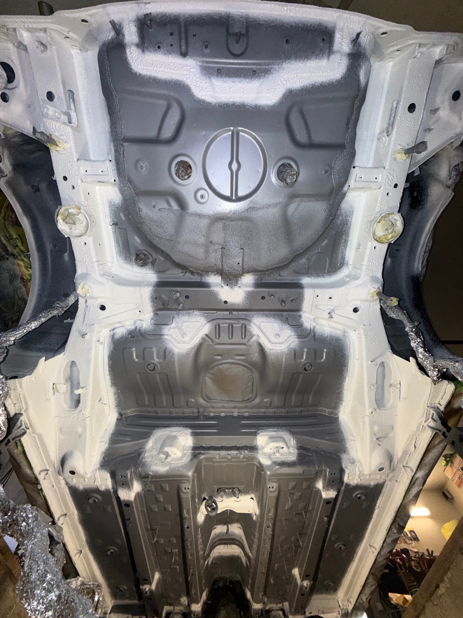 Podwozie samochodu po zabezpieczeniu antykorozyjnym, widok od spodu. Elementy w kolorze białym i szarym, zabezpieczone folią fragmenty nadwozia.