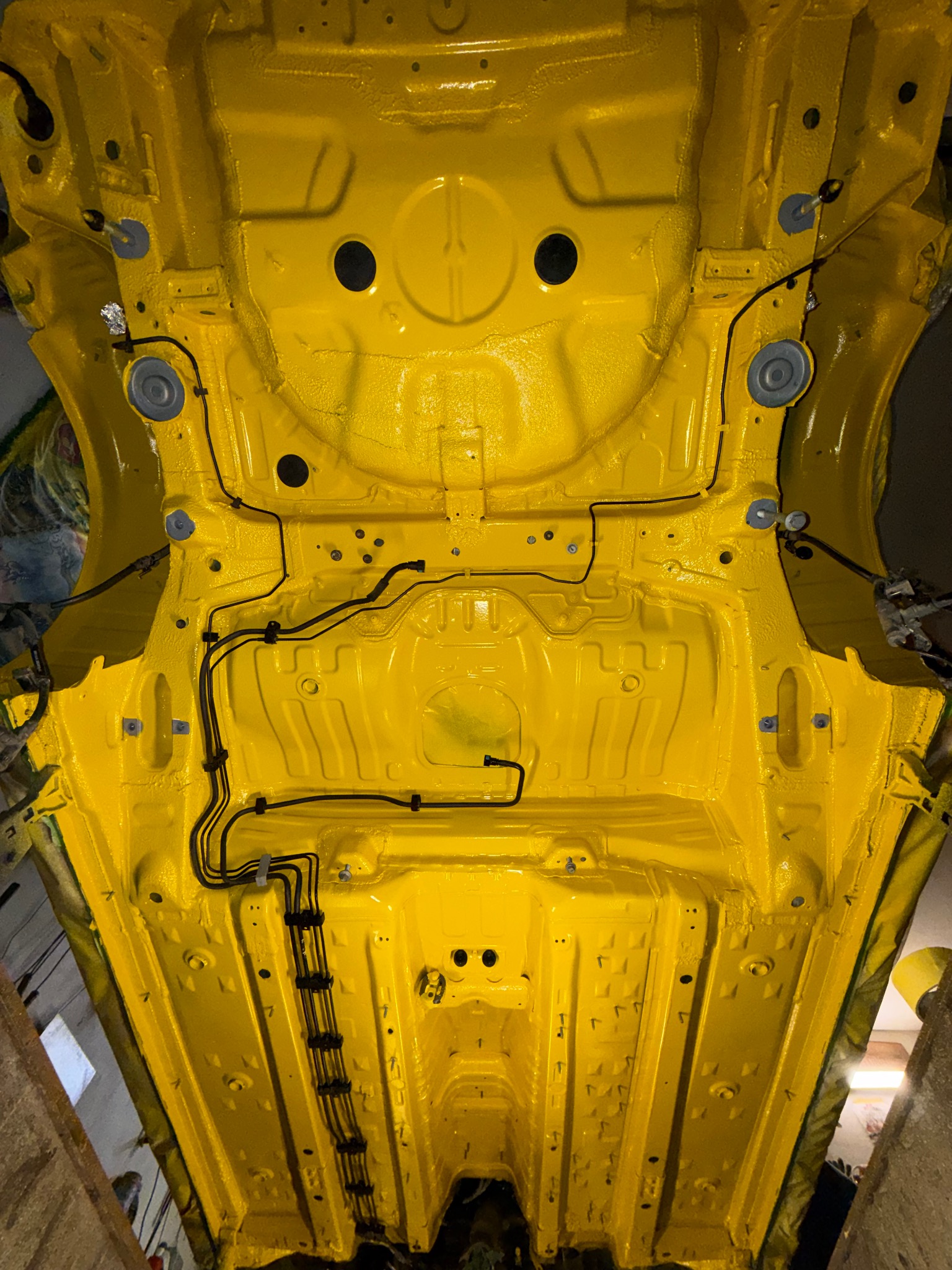 Żółte, świeżo pomalowane podwozie samochodu z widocznymi przewodami i elementami konstrukcyjnymi. Nowa powłoka antykorozyjna. Ujęcie od dołu, w warsztacie.