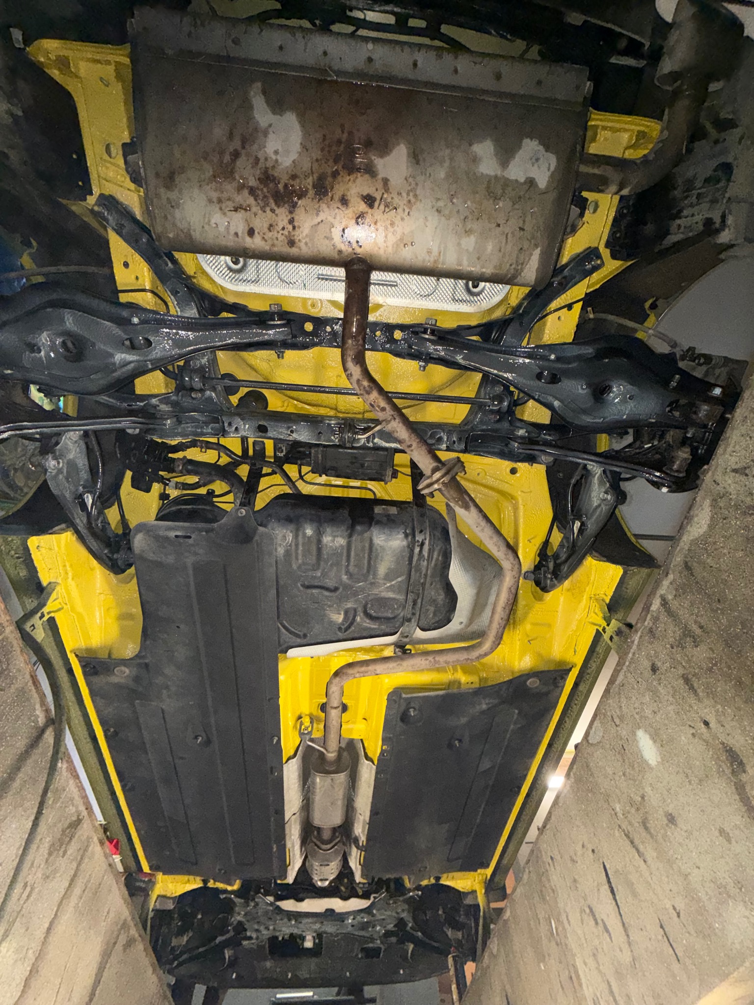 Żółte podwozie auta po konserwacji antykorozyjnej, widok od spodu. Elementy zawieszenia i układu wydechowego w kolorze czarnym. Widoczne ślady po konserwacji.