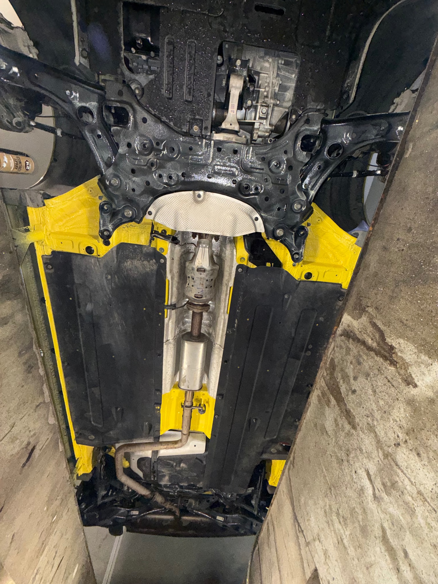 Żółta powłoka antykorozyjna podwozia samochodu na podnośniku, widok od spodu. Elementy zawieszenia w kolorze czarnym. Widoczne fragmenty układu wydechowego.