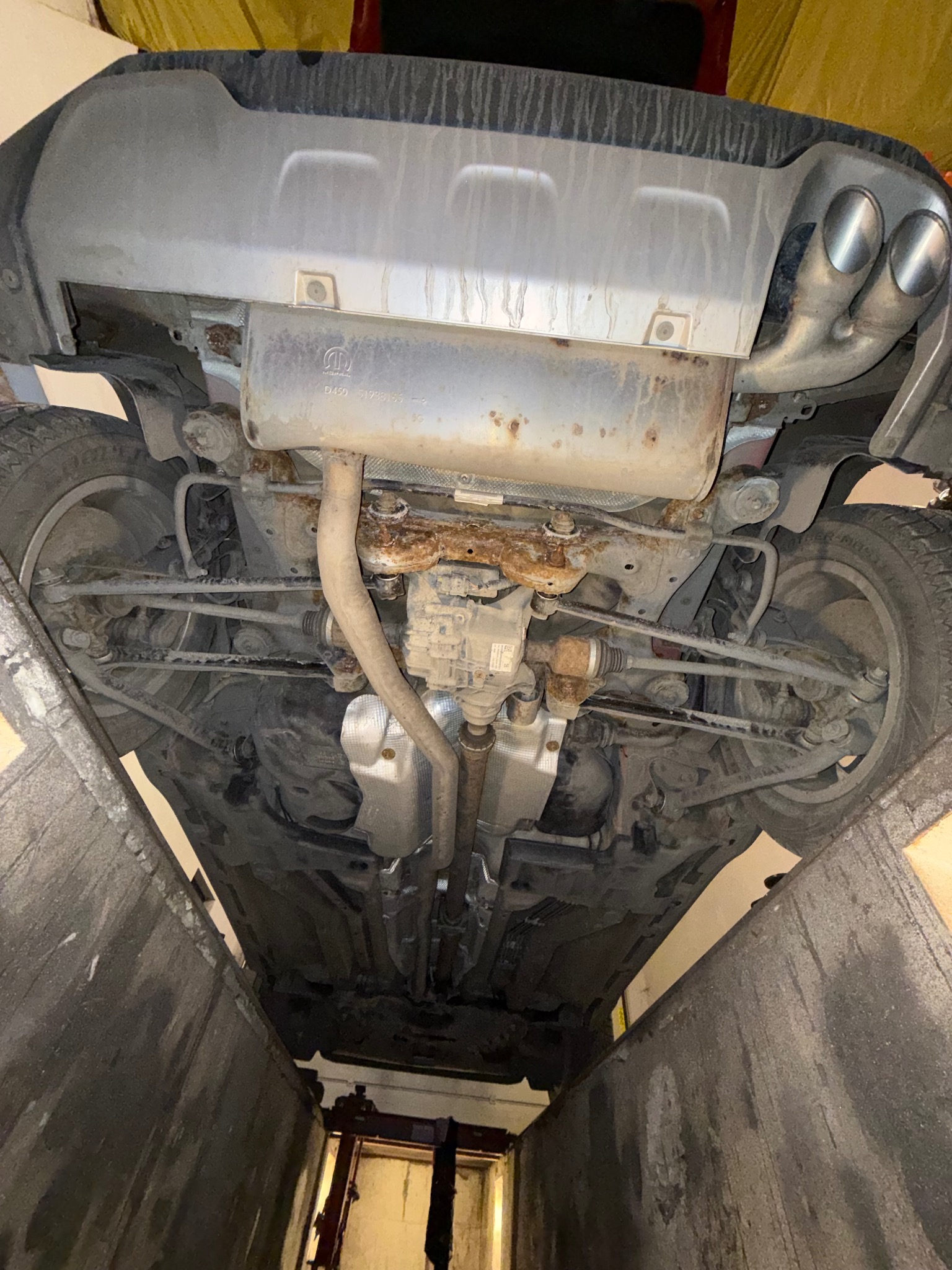 Widok od dołu na podwozie samochodu na podnośniku, z widocznymi elementami układu wydechowego i zawieszenia. Widoczne ślady korozji na elementach metalowych.