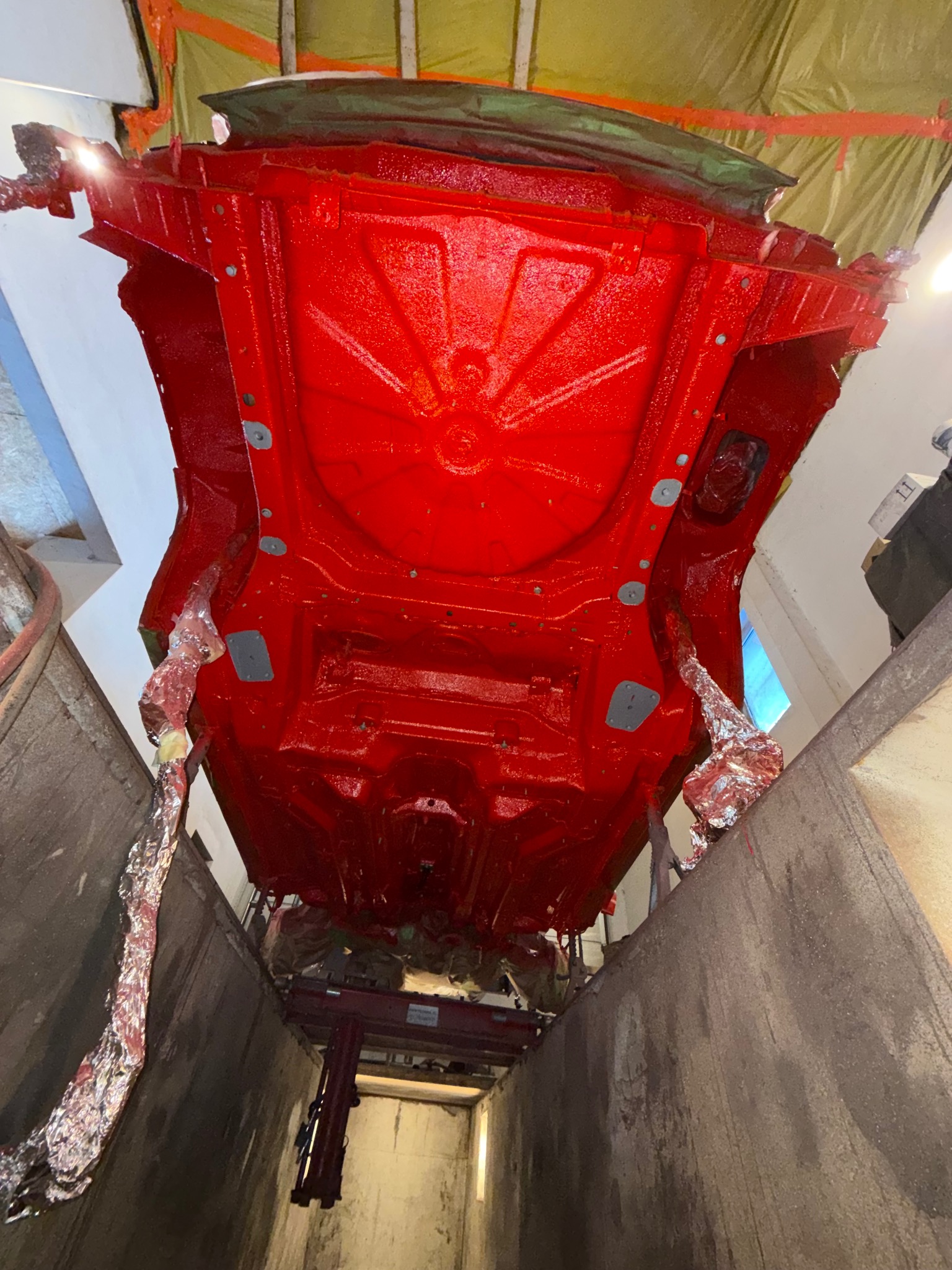 Czerwone podwozie samochodu, zabezpieczone folią, zawieszone w lakierni. Widok z dołu, ukazujący detale konstrukcji po nałożeniu warstwy ochronnej.