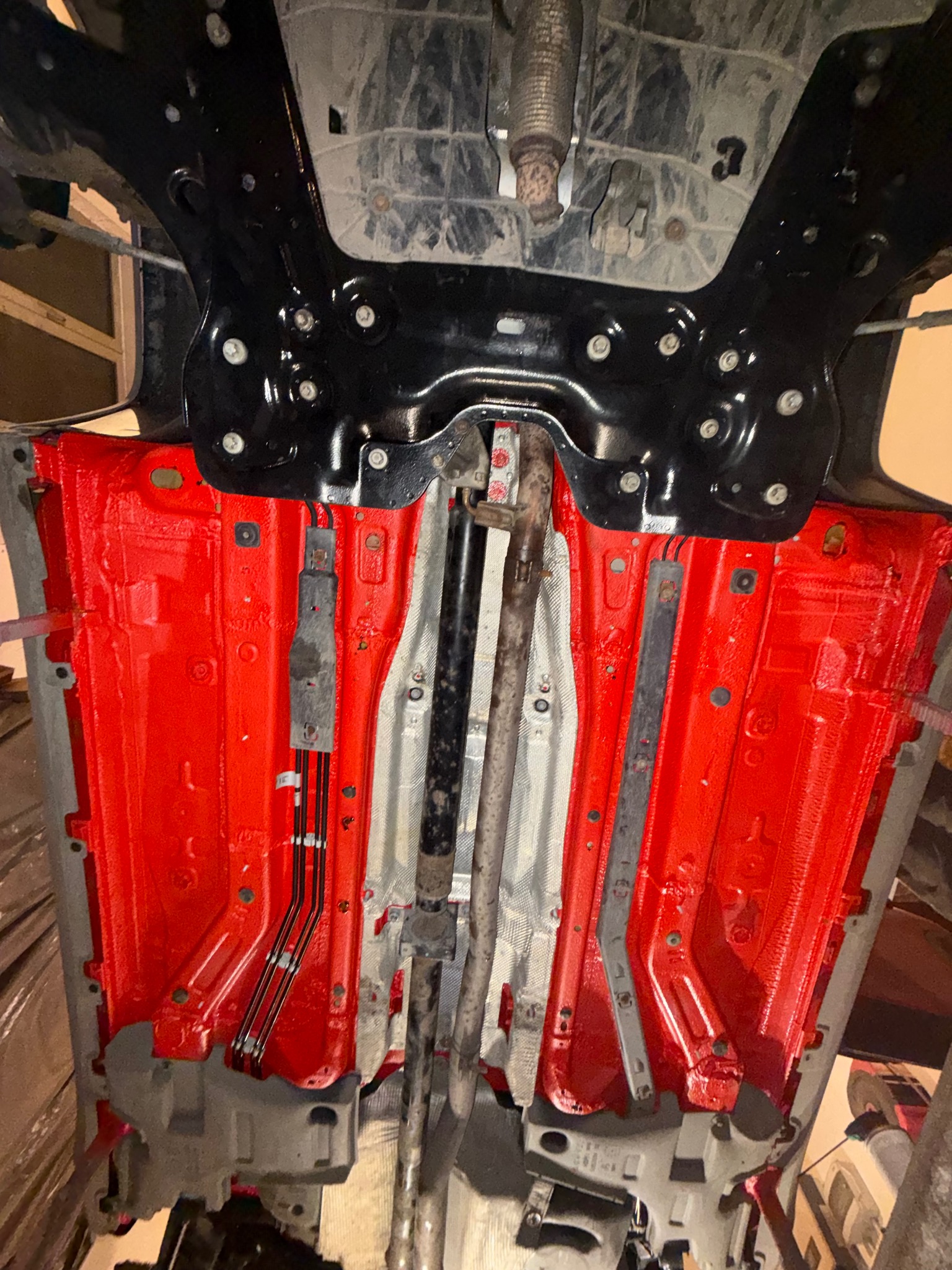 Czerwona, zabezpieczona antykorozyjnie podłoga samochodu na podnośniku. Widoczne elementy zawieszenia, układu wydechowego i przewody. Ujęcie od dołu.