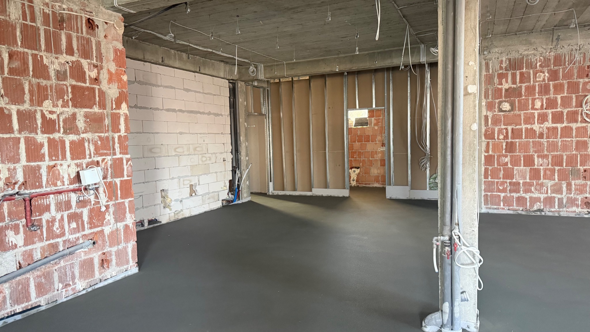 Szara, gładka wylewka podłogowa w surowym wnętrzu z instalacjami i ścianami z cegły oraz bloczków. Widoczne przewody i rury. Surowy stan deweloperski.