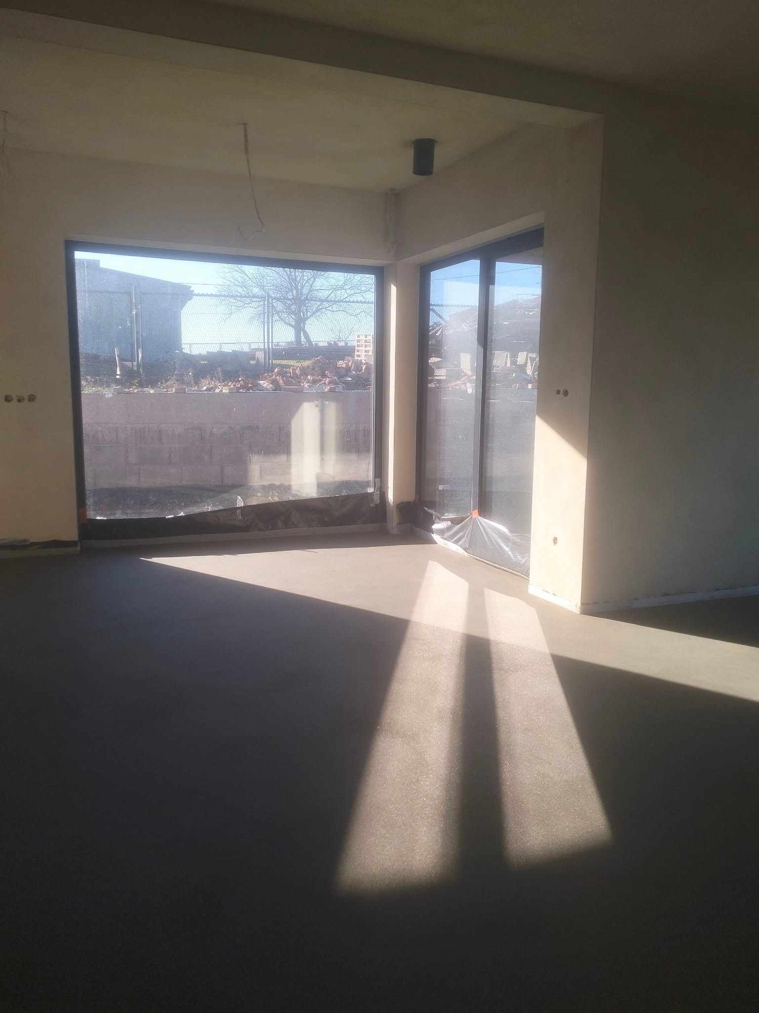 Świeżo wylana, gładka posadzka w jasnym wnętrzu z dużymi oknami. Promienie słoneczne tworzą geometryczne wzory na powierzchni. Widoczne przygotowania do dalszych prac.