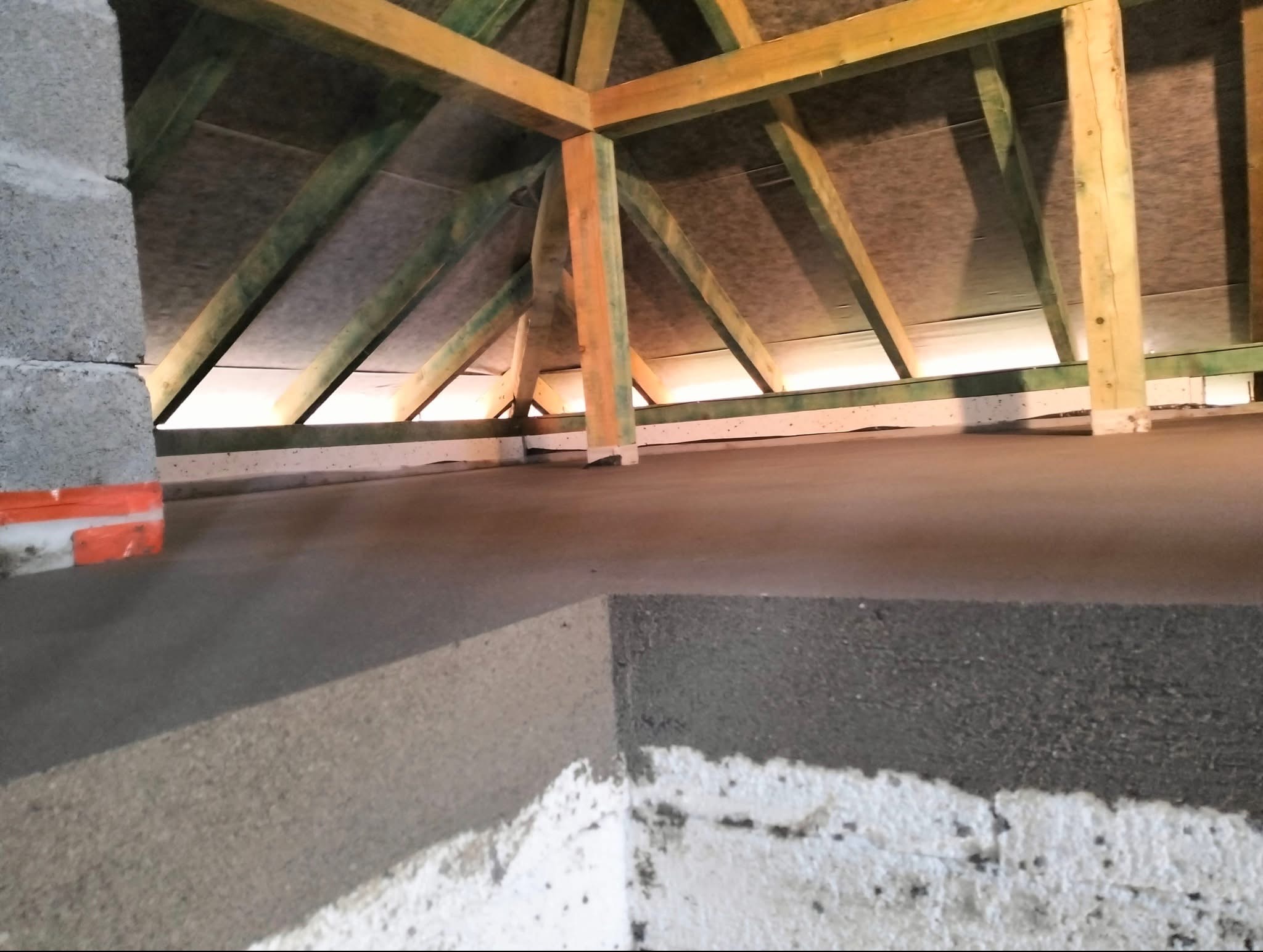 Wyrównana, szara posadzka jastrychowa na poddaszu z drewnianą konstrukcją dachu. Widoczny fragment ściany z cegieł i izolacja. Surowy stan wykończenia.