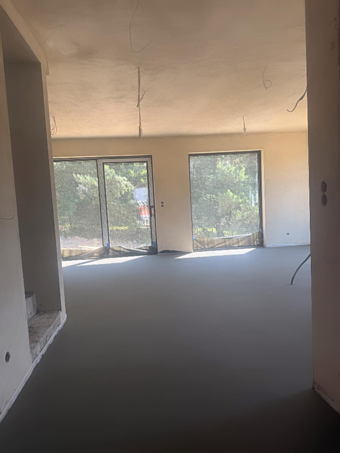 Wnętrze z wylaną, gładką posadzką w odcieniach szarości, widoczne okna z widokiem na zieleń. Minimalistyczny styl, surowe wykończenie ścian i sufitu.